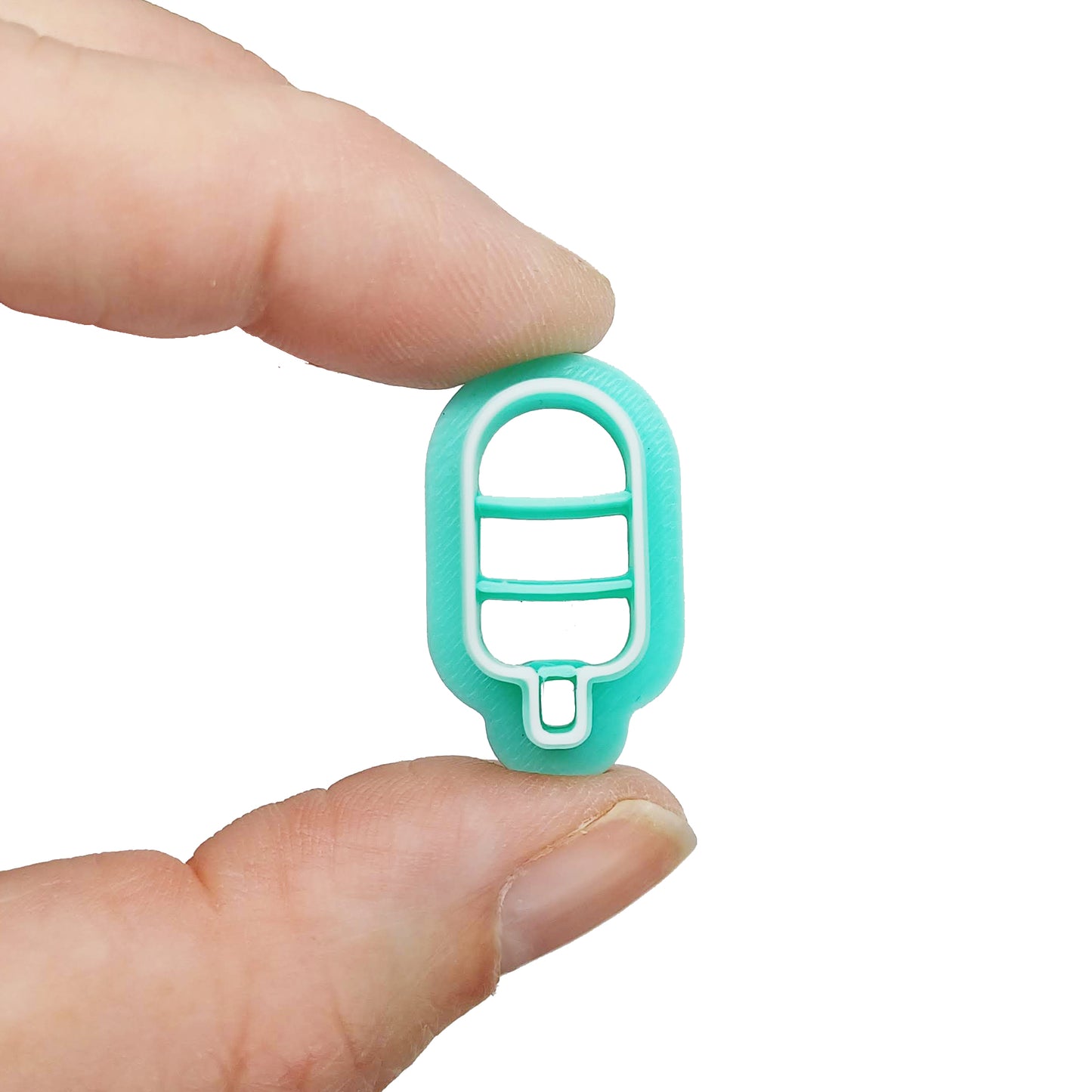 Turquoise clay cutter with an ice pop or ice cream shape, with white accents held between two fingers on a white background