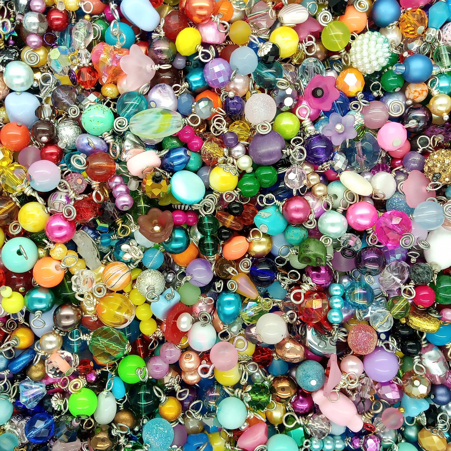 A dense, close-up pile of colorful craft beads dangle charms with silver metal loops, featuring various shapes, sizes, and glossy finishes in a rainbow of colors.