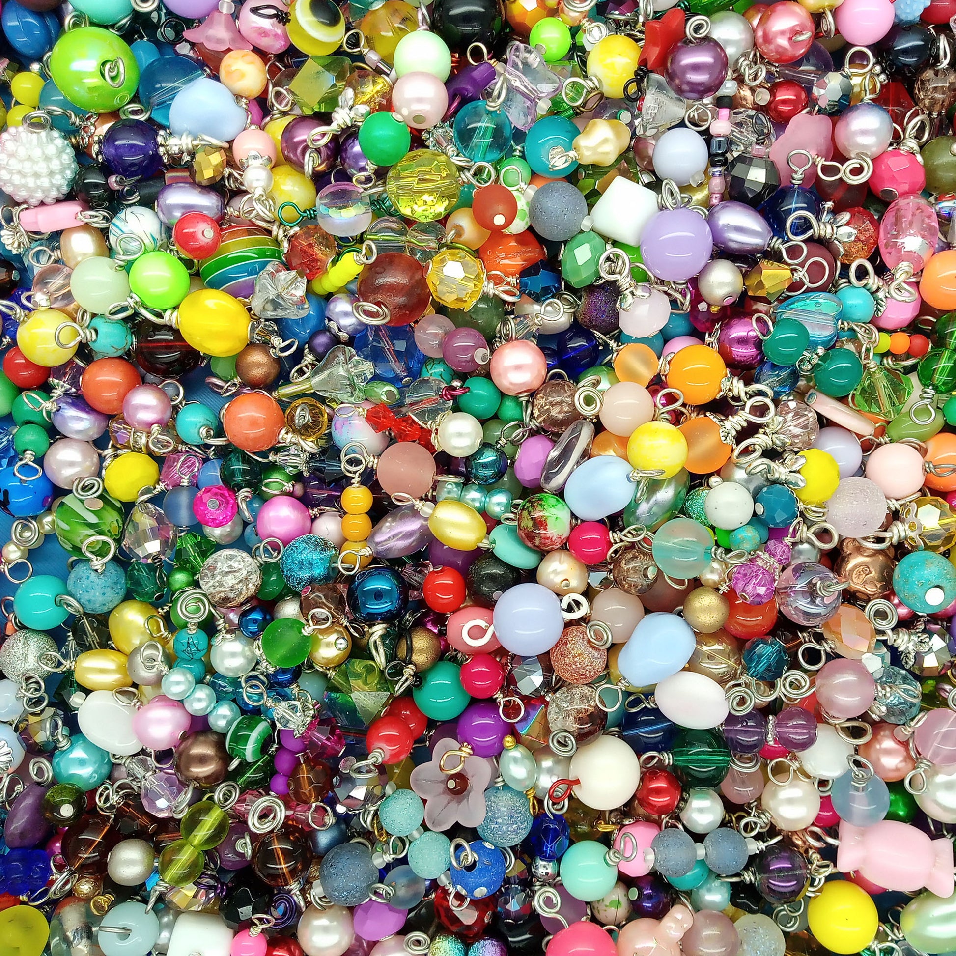 A dense, close-up pile of colorful craft beads dangle charms with silver metal loops, featuring various shapes, sizes, and glossy finishes in a rainbow of colors.