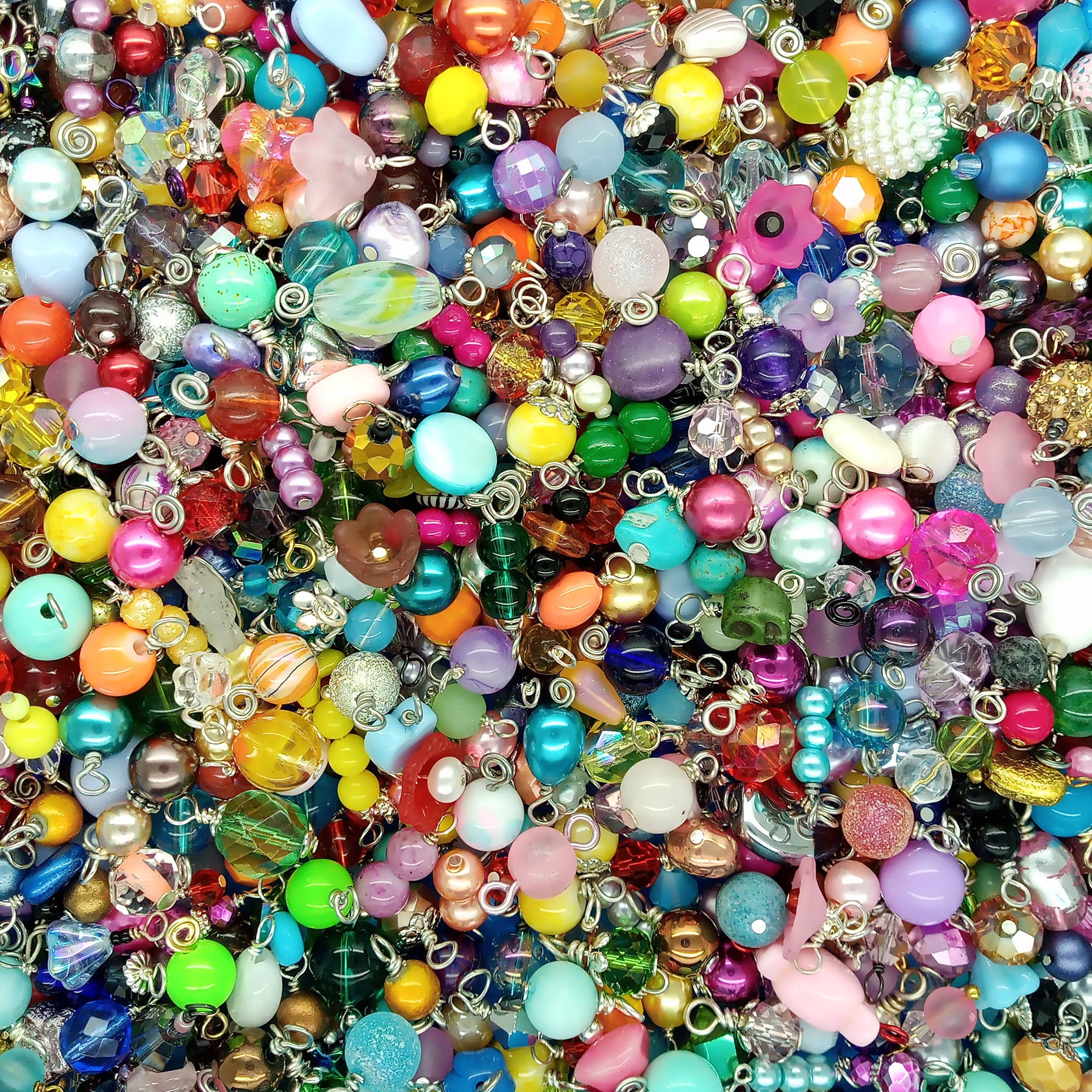 A dense, close-up pile of colorful craft beads dangle charms with silver metal loops, featuring various shapes, sizes, and glossy finishes in a rainbow of colors.