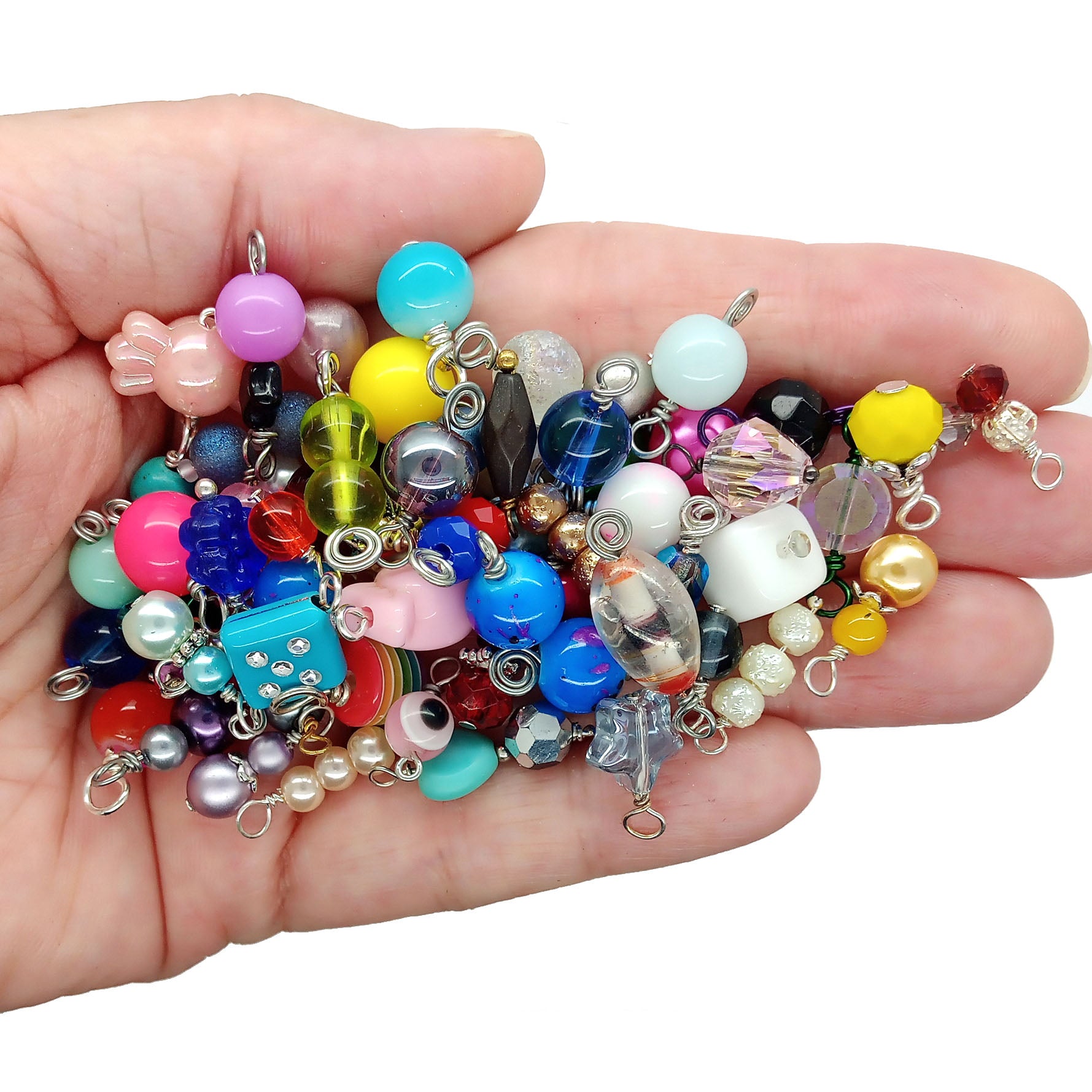 A hand holding a colorful assortment of craft beads charms with silver loops, shown against a plain white background.