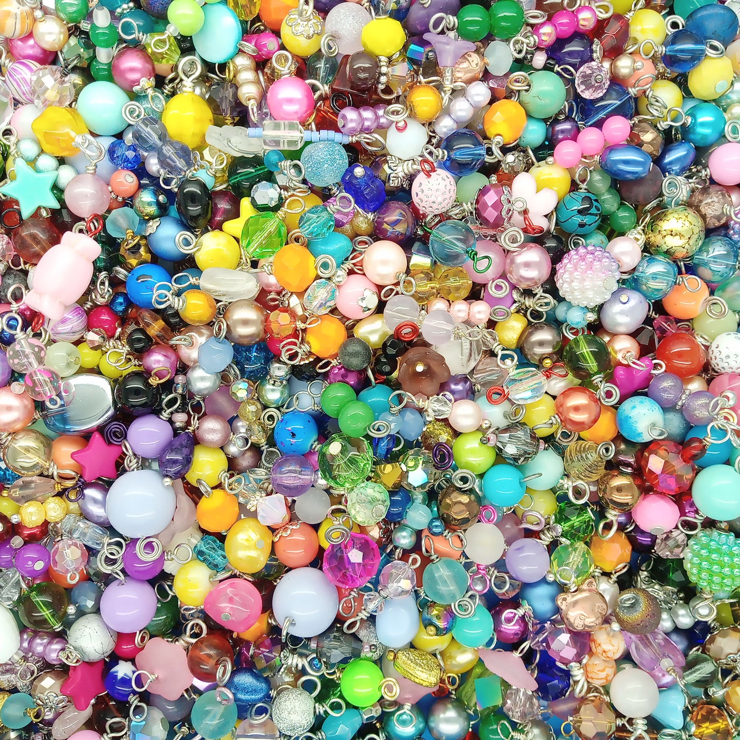 A dense, close-up pile of colorful craft beads dangle charms with silver metal loops, featuring various shapes, sizes, and glossy finishes in a rainbow of colors.