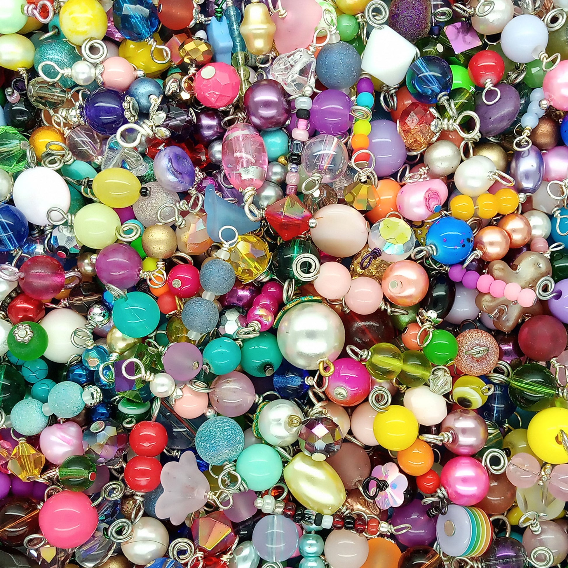A dense, close-up pile of colorful glass bead charms and jewelry findings with silver metal loops, featuring various shapes, sizes, and colors including pink, purple, blue, green, yellow, and white.