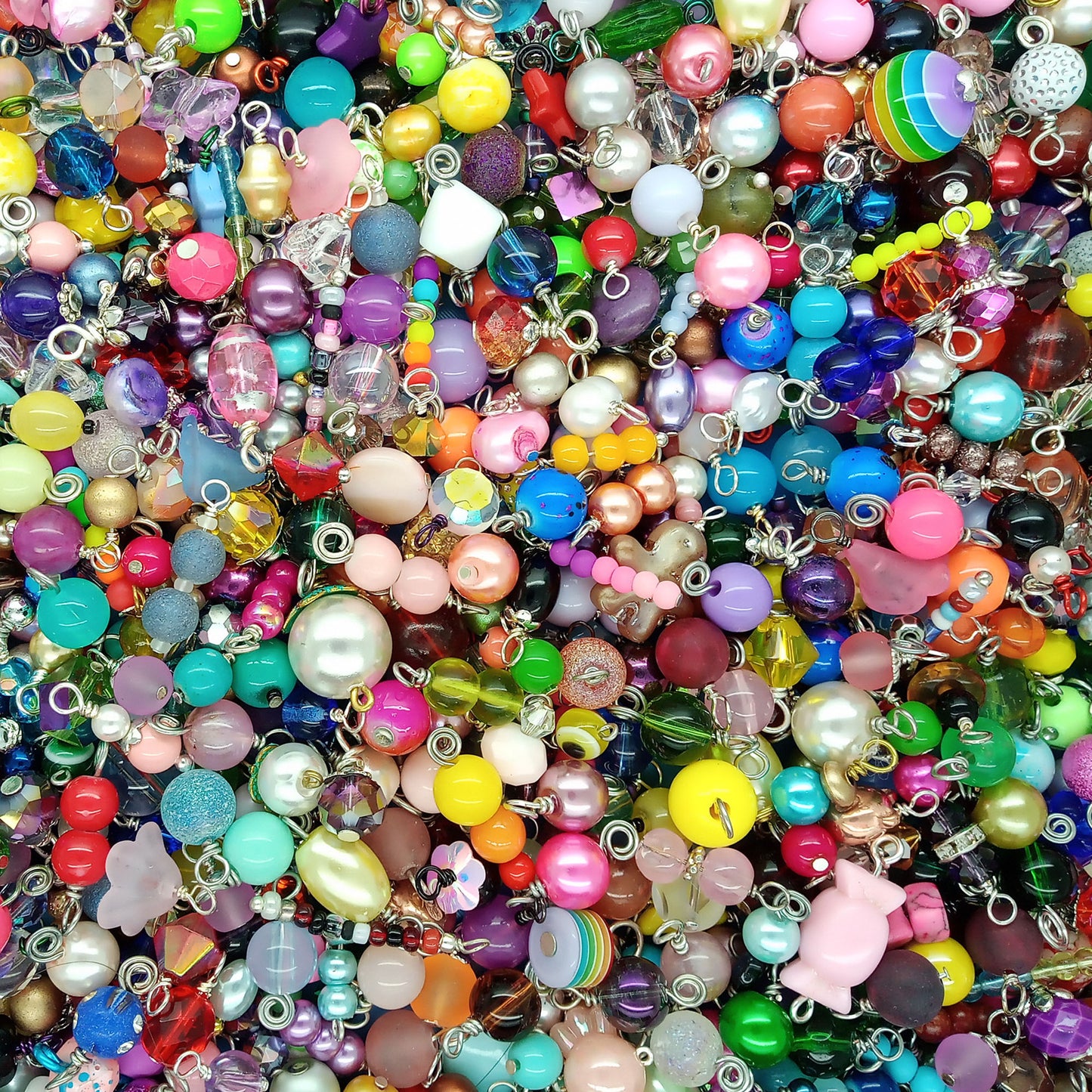 A dense, close-up pile of colorful craft beads dangle charms with silver metal loops, featuring various shapes, sizes, and glossy finishes in a rainbow of colors.